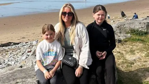 Family photo A young white girl wearing a t-shirt saying hello, her mother and her older sister sit on a rock at the edge of the beach. The mother, Lucy, has long blonde hair and wears black trousers, a plain tshirt and a white light jacket and black handbag over it. She is wearing sunglasses. Lola to her right is aged about 11, is wearing black trousers and sport top. all three are smiling at the camera. It's a sunny day. A few people sit on the beach behind them.