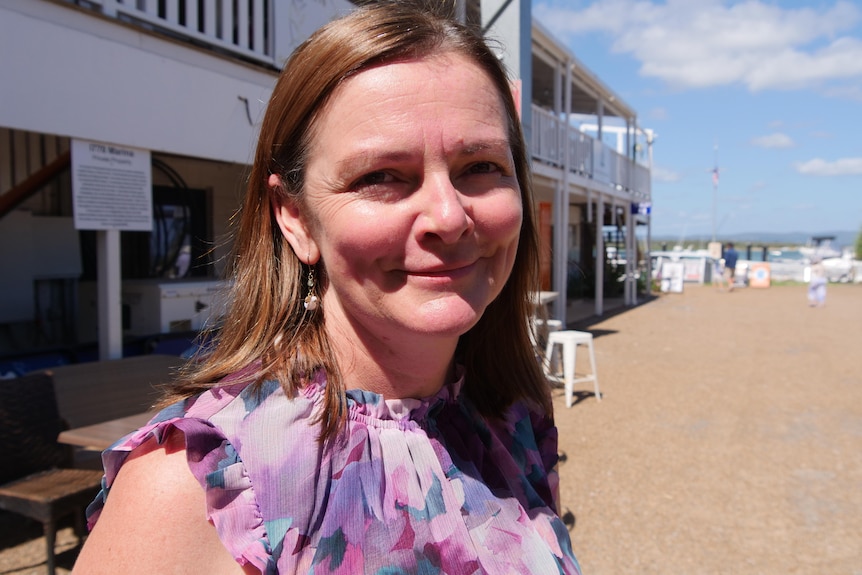 Woman smiling gently at camera, wearing a pink and purple top, with local shops in the background.
