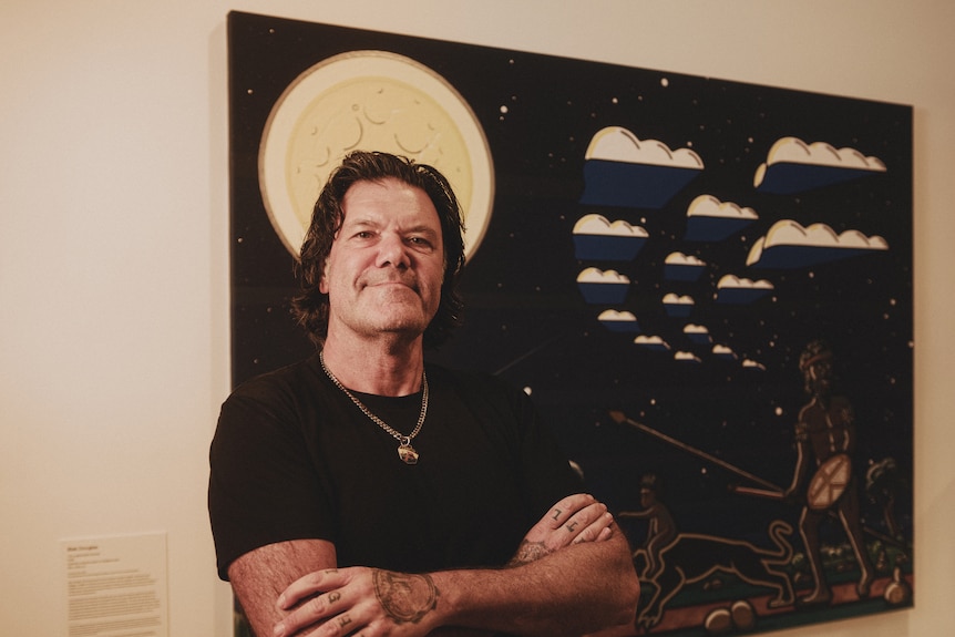 An Aboriginal man with his arms crossed, standing in front of his painting, ‘The Original Blak Panther’.