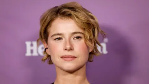 REUTERS/Caroline Brehman A close-up of Jessie Buckley attending the 2026 BAFTA Tea Party in Los Angeles.  She has short, wavy blonde hair and is pictured in front of a purple background. 