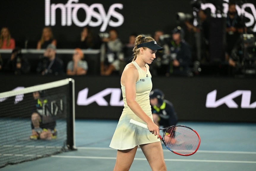 A tennis player in cream colours and black cap looks down while holding racquet