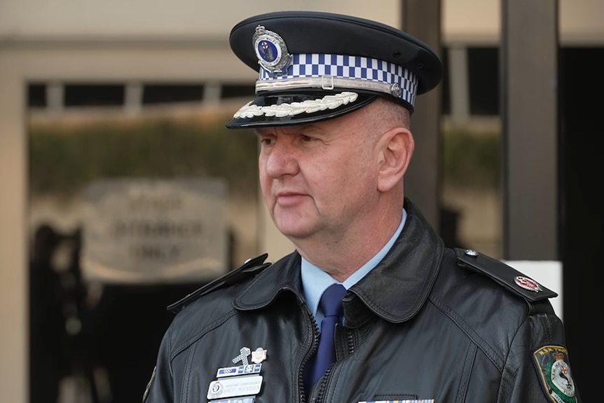 A middleaged police officer in uniform speaks into a set of microphones outside a brick building.