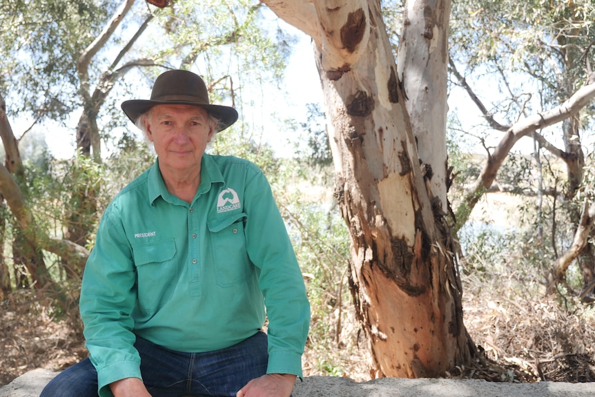 Simon wears a long sleeved Landcare shirt and a wide brim hat, sitting on a bench in front of a tree at the Nature Park