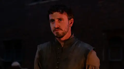 Universal Pictures/Focus Features Paul Mescal has short dark hair and a light beard. He is wearing a grey waistcoat and lighter grey shirt. He is illuminated by a yellow light with the background in darkness. 