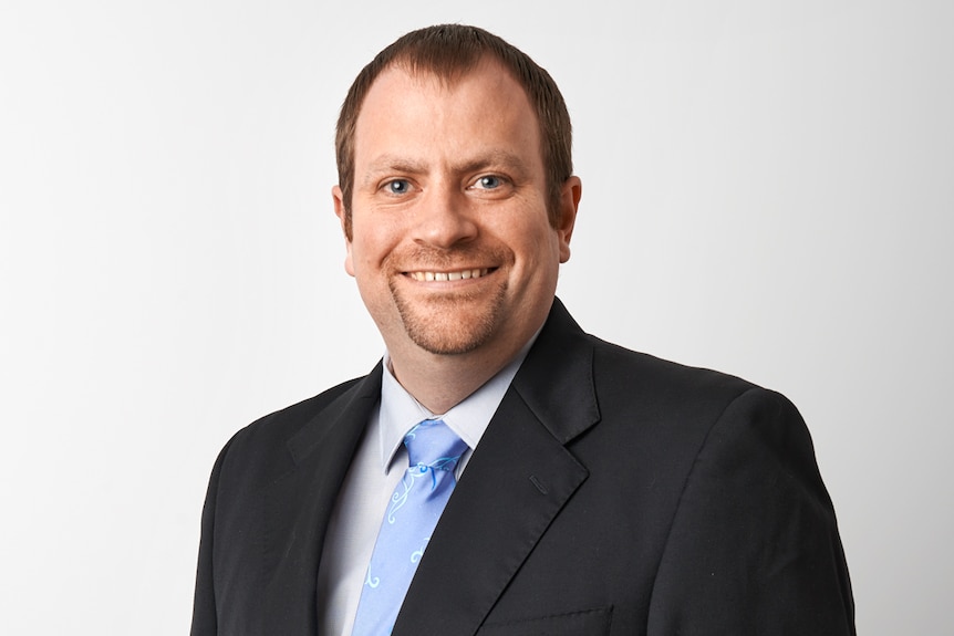 A middle-aged man with short hair and a goatee wearing a suit with a blue tie smiling