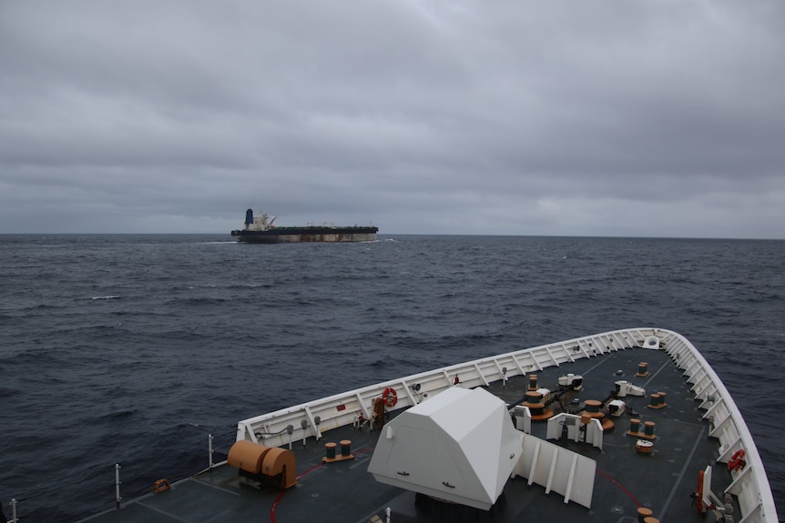 An oil tanker sits on a grey ocean against a grey sky with the front of another boat in the foreground