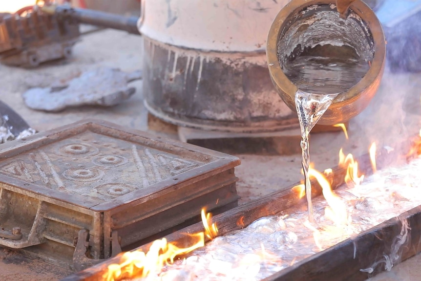 Molten metal is poured from a jug onto a flaming and bubbling mould.