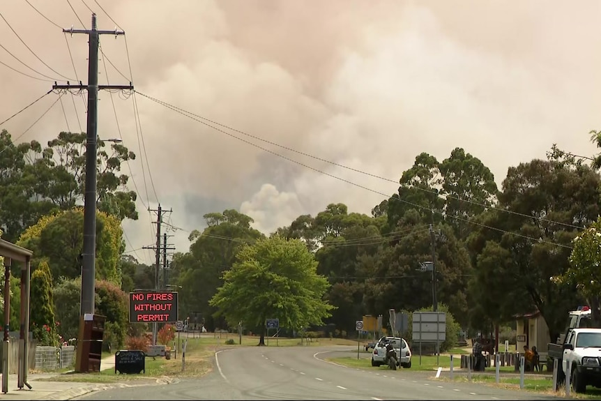 Thick smoke billows into the sky at the end of a street in a country town where an electronic sign says no fires are allowed.