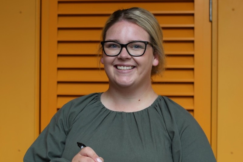 A young woman with blonde hair holds a pen and notepad, smiling.