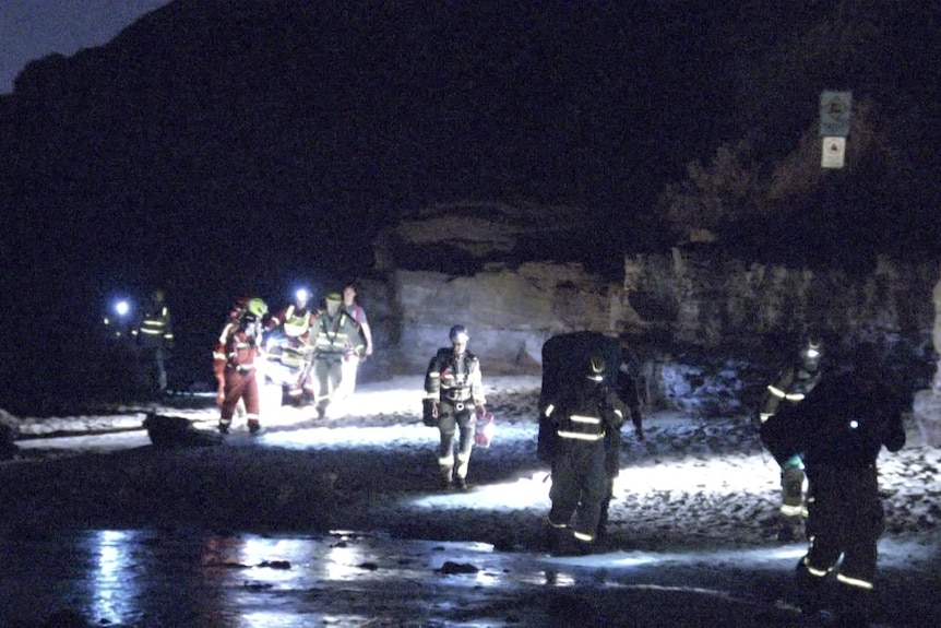 People dressed in overalls and carrying torches guide a stretcher along the sand beside the water at night.