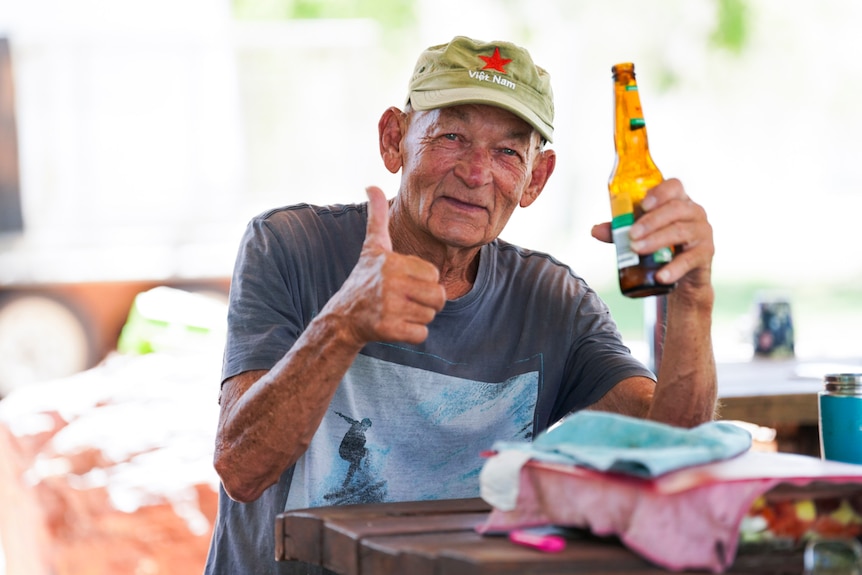 A man in a grey shirt wears a green cap and holds a beer and gives the thumbs up.