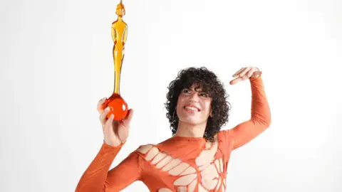 PA Media Jacob Alon is looking up at a Brit Award which they are holding in their hand. They wear a orange ripped top and have brown curly hair. 