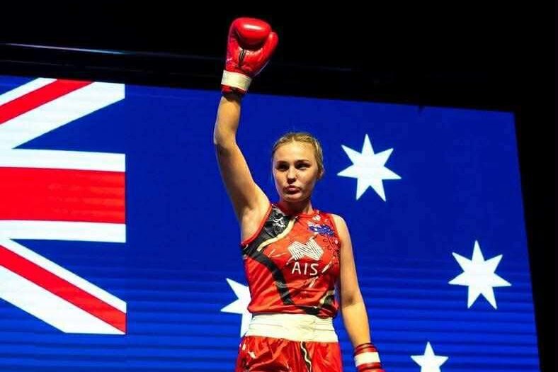 A woman wearing boxing gloves raising her hand with the Australian flag in the background.