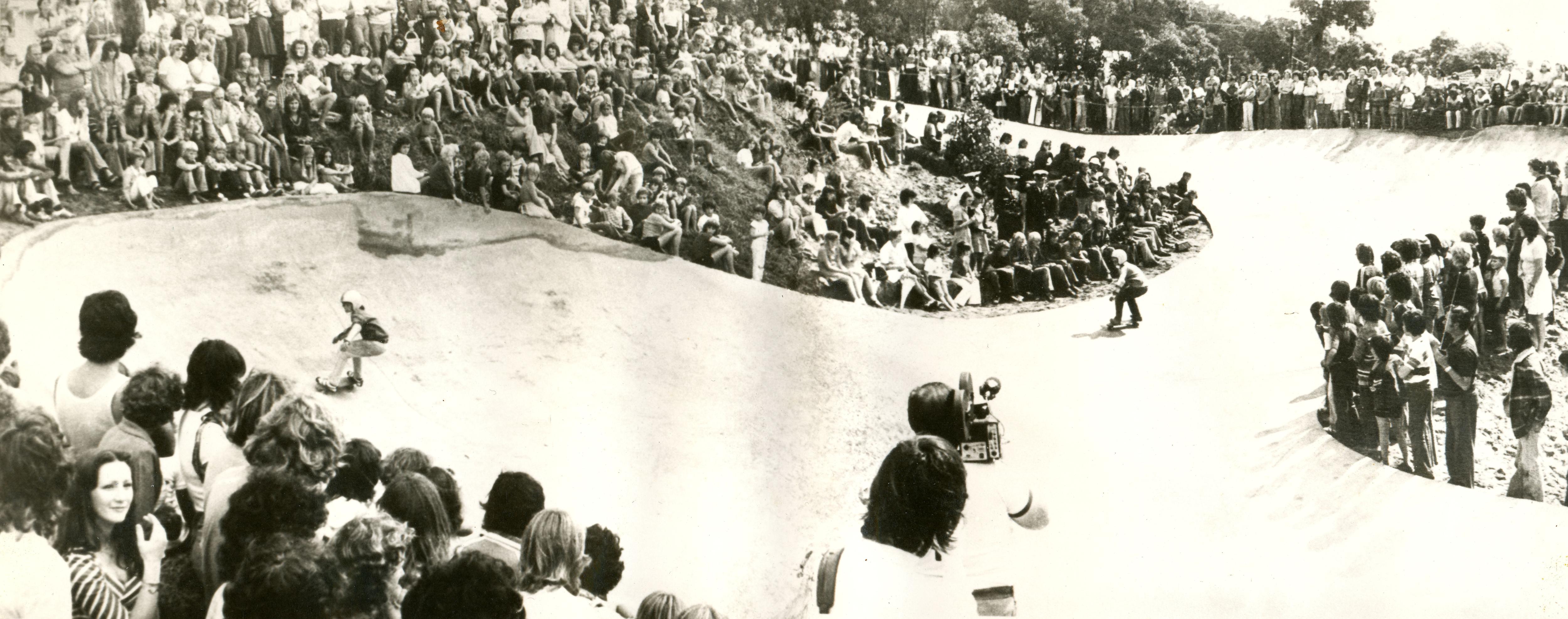 A black and white photograph of crowds watching people skate at a skate park. 