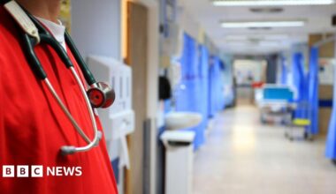 A stock image of a hospital ward. The image shows an empty ward with several bays with blue curtains, which are blurred in the background. In the forefront of the image is a man in bright red scrubs with a stethoscope around his neck.