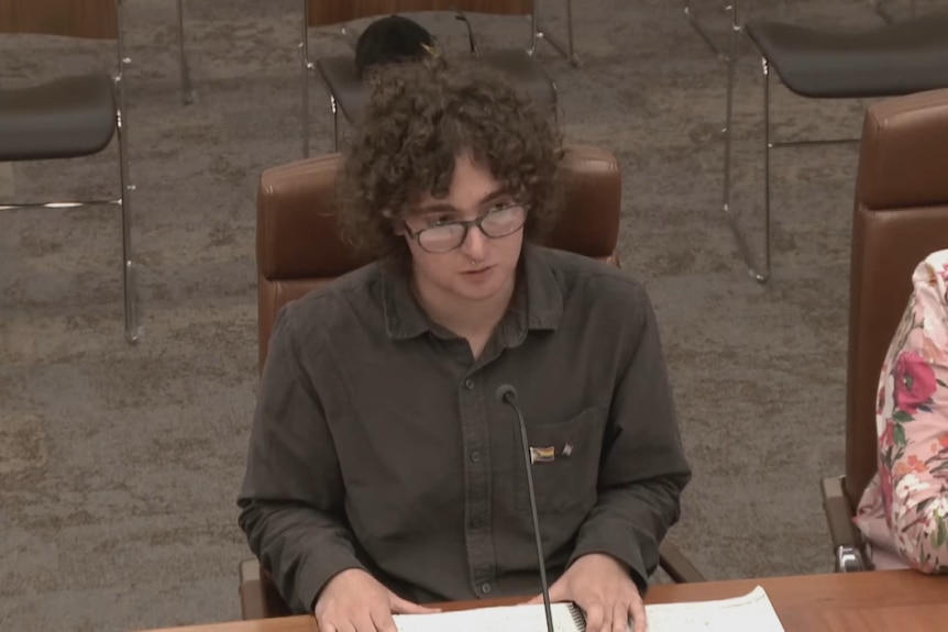 A man with glasses and a dark shirt sits at a wooden desk in front of a microphone.