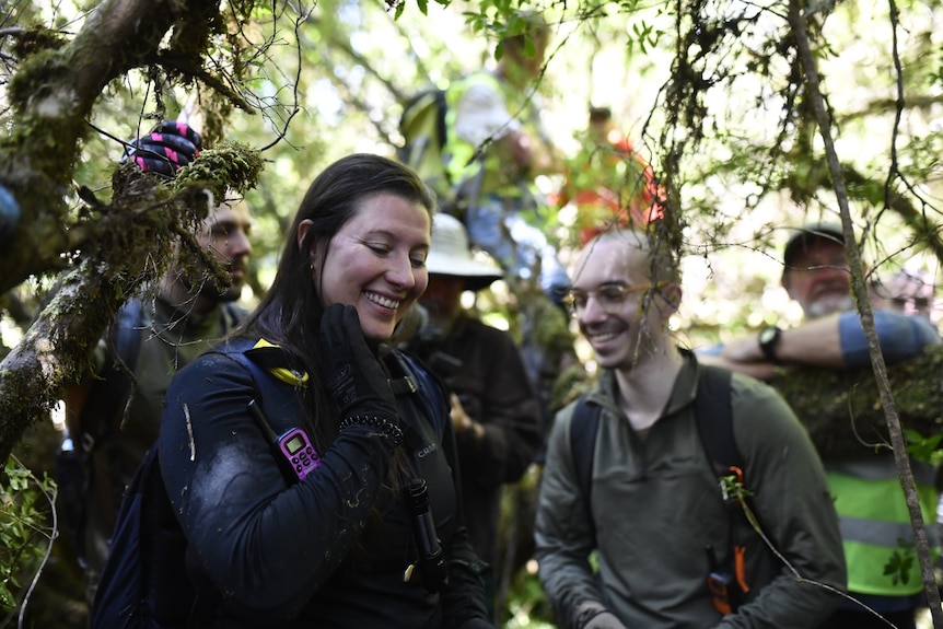 Rachil Disbechl stands amongst trees with other people and smiles as she looks down.