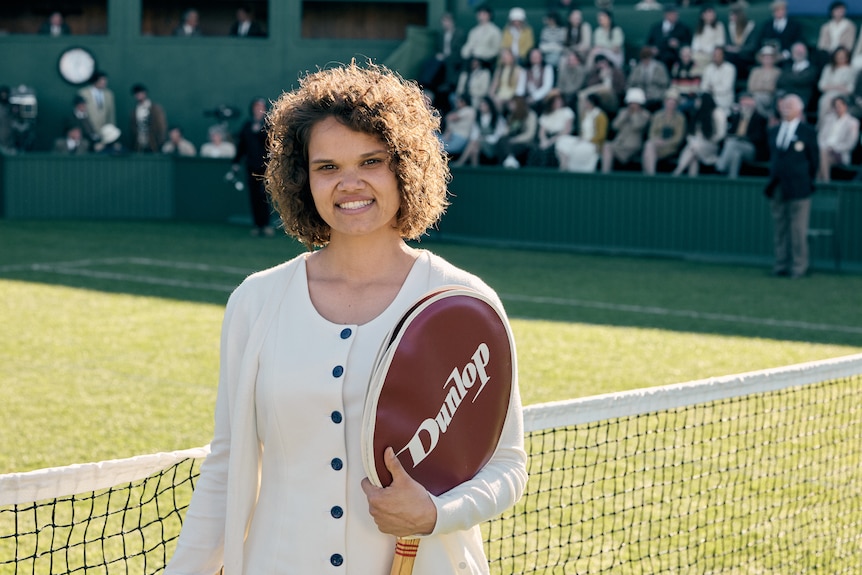 Holding a tennis racket, Lila smiles from the tennis court.