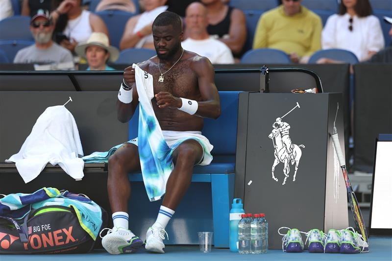 tennis player frances tiafoe sits courtside with his shirt off, water bottles at his feet