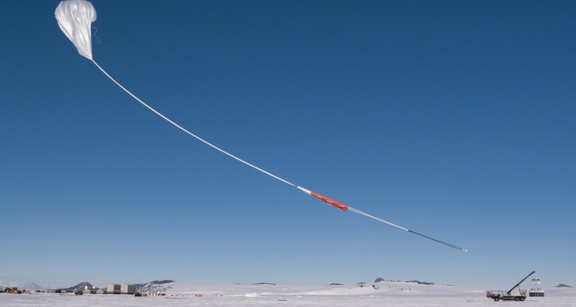A white scientific balloon rises into the bright blue sky above snowy Antarctica. The sky and the balloon take up most of the image, with the ground only being a small white stripe at the bottom of the photo. The balloon has a long white "tail" with orange and black portions.