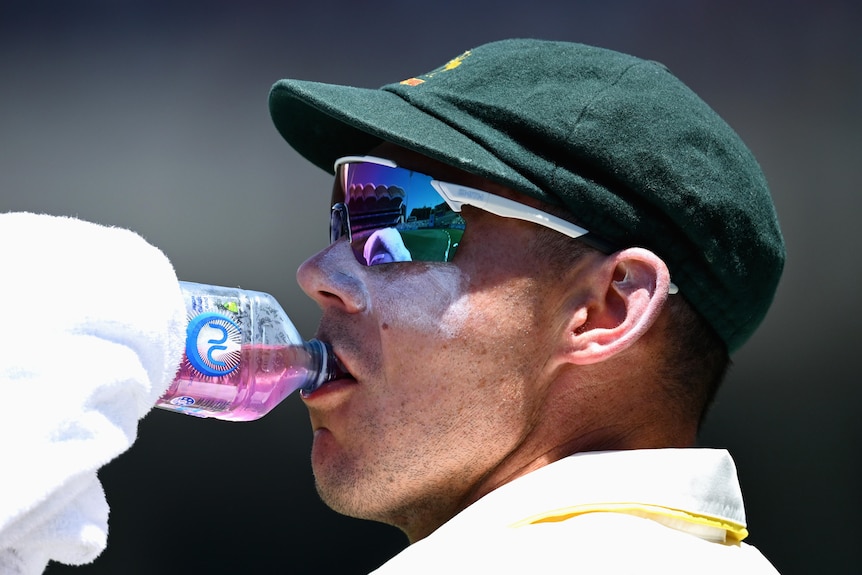 Australia bowler Scott Boland drinks from a water bottle while wearing sunglasses and a cap.