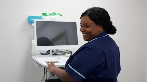 Luca Felice/Milton Keynes University Hospital NHS Foundation Trust A nurse in a navy uniform operates a computer workstation inside a clinical room, with medical equipment and cables visible on the unit. The nurse is smiling.