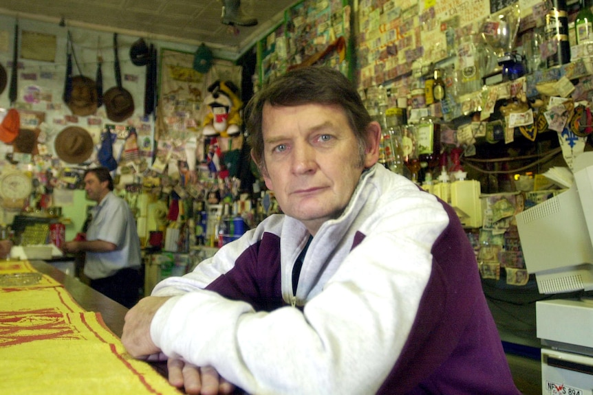 A man with blue eyes and dark hair, standing behind the bar inside an outback hotel.