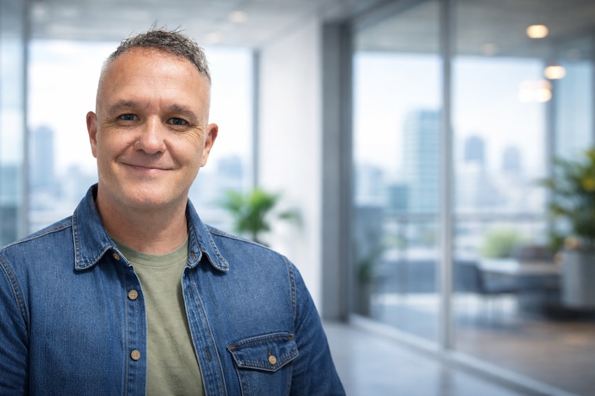 A man smiles at the camera in a city office wearing a green shirt with a blue long shirt over the top