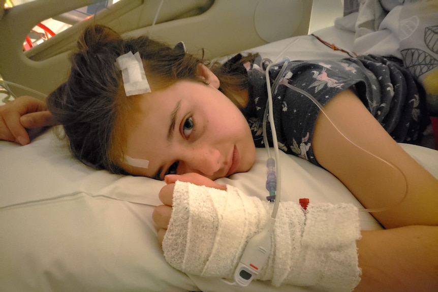 Abbey Barrett lies on a hospital bed with her wrist and hand bandaged, and with two small bandages on her head.