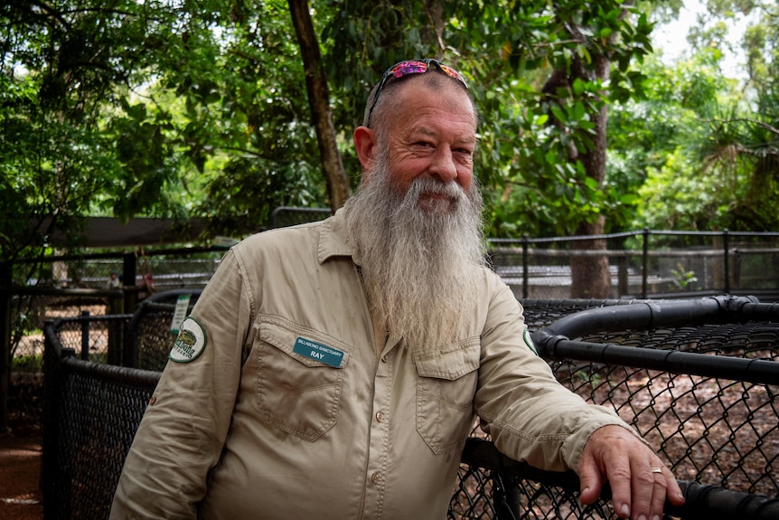 A man in a wildlife uniform leans on the fence of a croc enclosure, smiling