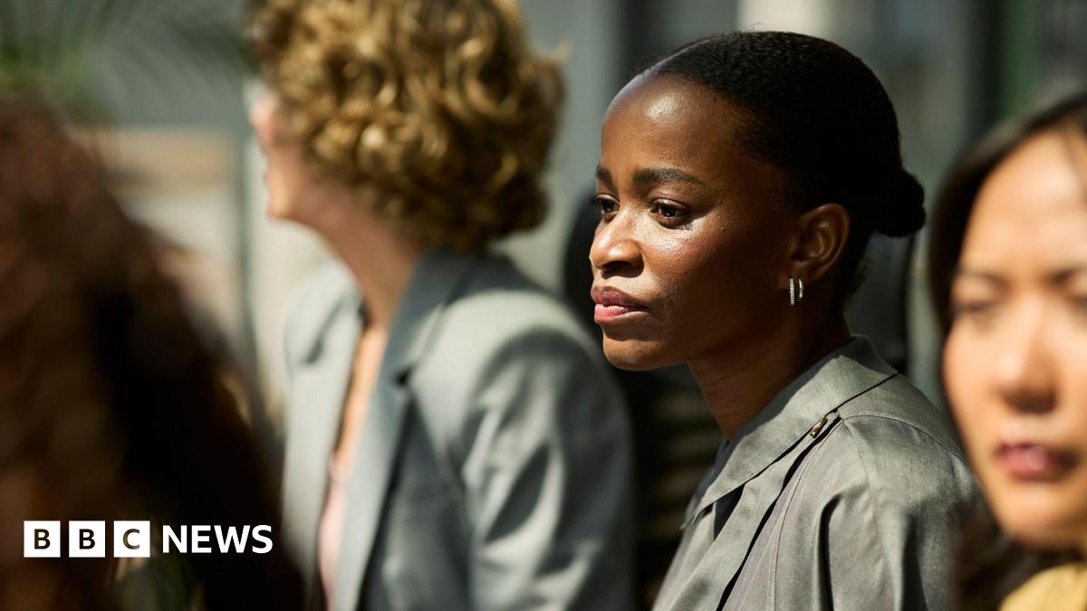 A young black woman with her hair tied back in a bun gazing with a concerned look on her face during a meeting with other female colleagues blurred out of focus around her