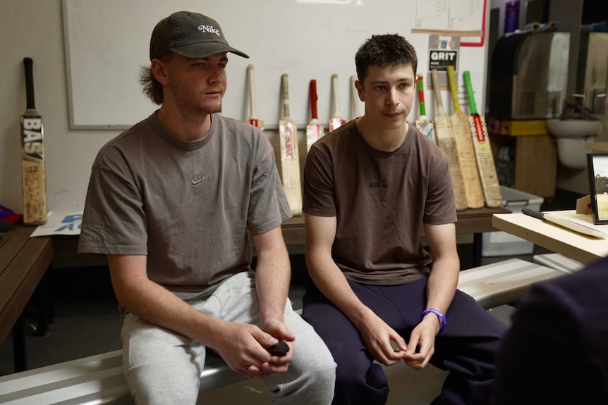 Two young men sitting on a bench. Cricket bats are lined up against a wall behind them.