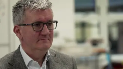 Matthew Trainer, is seen wearing glasses and a jacket sits indoors, looking slightly to one side.