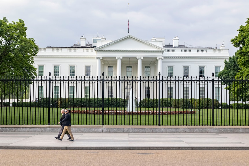 A picture of the White House taken from outside on the street, with two men walking by a metal fence.