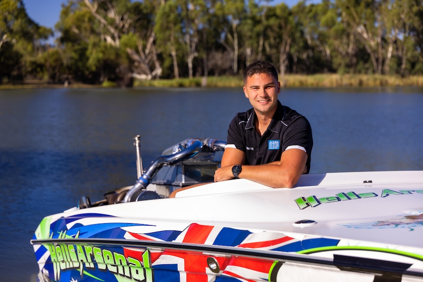 A man in a black polo near a river, leaning on a ski boat that says Hells Arsenal in green letters