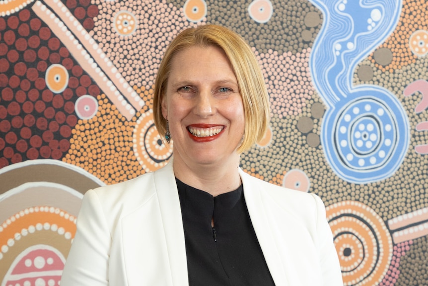 A blonde woman with a white blazer in front of an Indigenous painting.