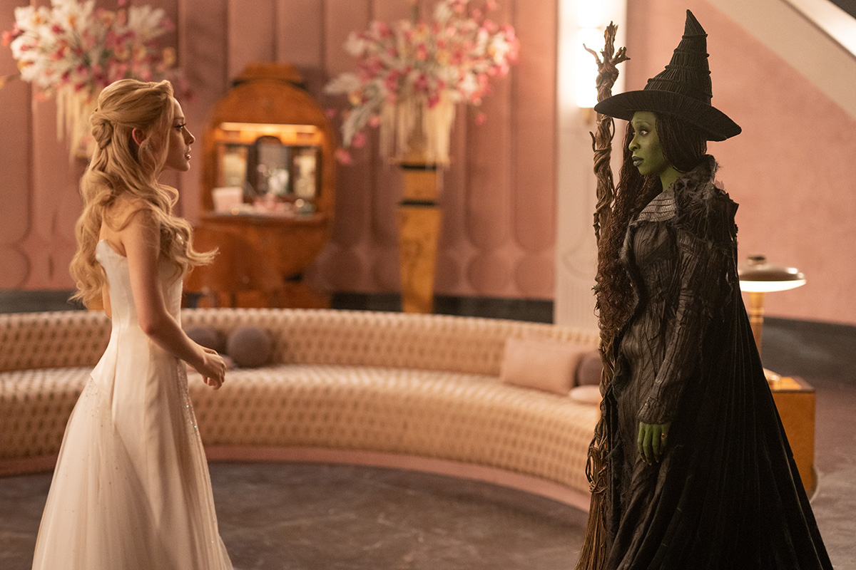 A blonde woman in a pink dress looks at a woman with green skin who wears a witch's hat and holds a broomstick.