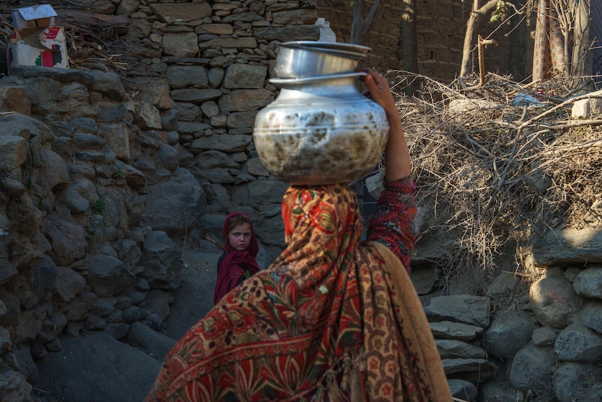 An Afghan woman balances a pot on her head while a child looks on.
