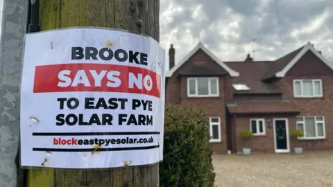 Jenny Kirk/BBC A close-up on a telegraph pole which has a sign stuck to it saying "Brooke Says No To East Pye Solar Farm"
