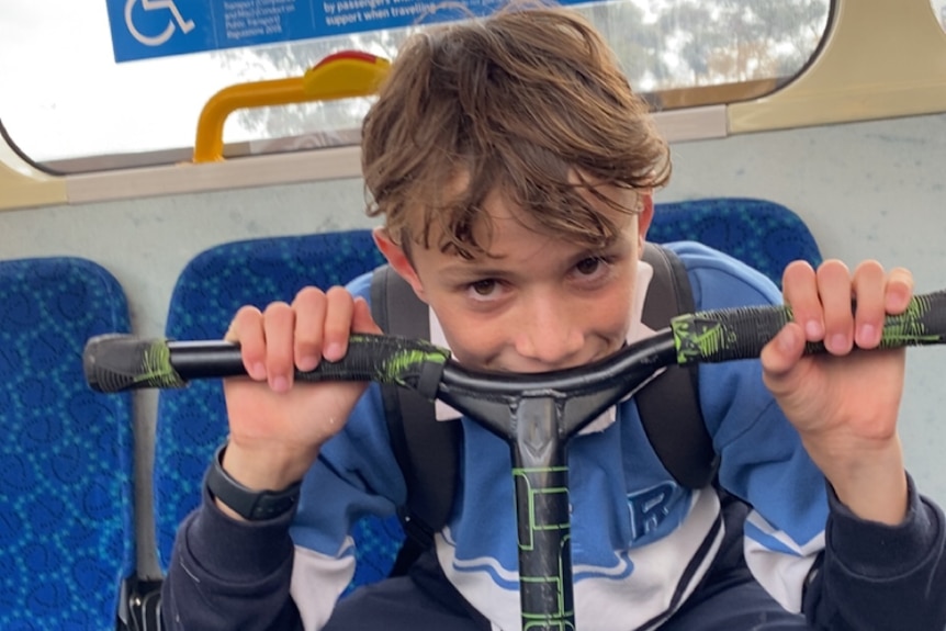 A boy leans on the handles of a scooter while travelling on a train or a tram.