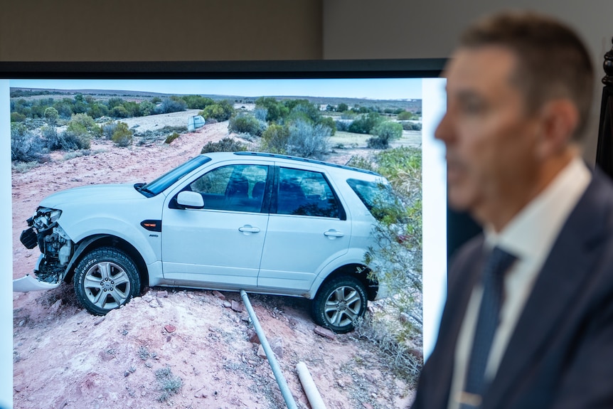 A photos of a white four wheel drive car with a damaged front end stuck on top of a mound of dirt on a projector. 