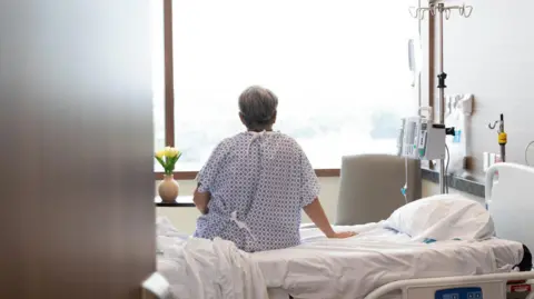 Getty Images A man sits on a hospital bed facing a window. His back is facing the camera.