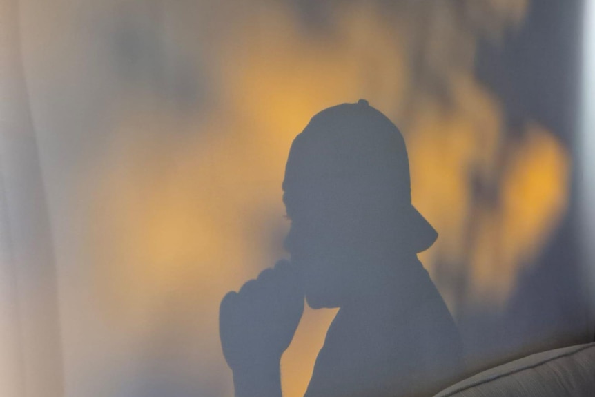A silhouette of a man, shadow on wall, wears cap backwards, hand is near mouth.
