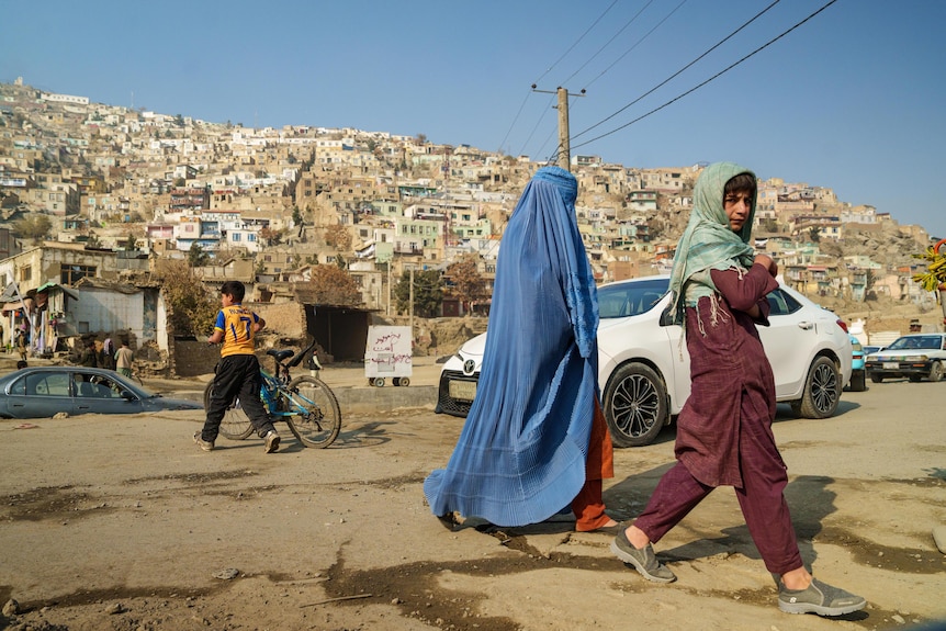 A woman wears a blue burka and a small boy near her looks at the camera, backdrop of Afghan homes on the hills 