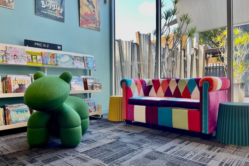 A kids' section at a library with a large green stuffed dinosaur, a colourful couch and books on shelves