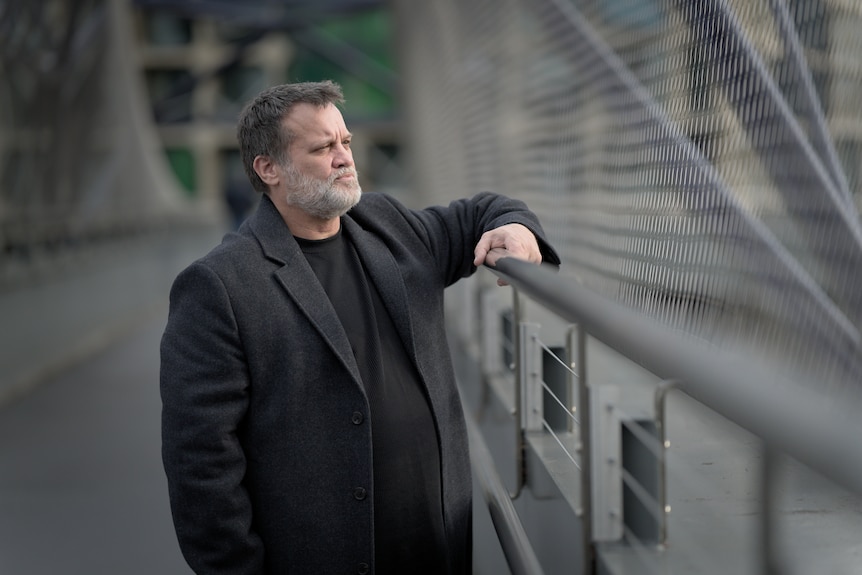 Nicholas Taylor  stands on a pedestrian bridge and leans on a metal railing.