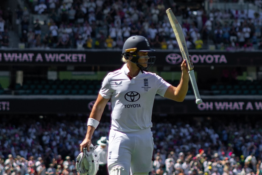 A cricketer leaves the field holding his bat high in the air with the crowd applauding him