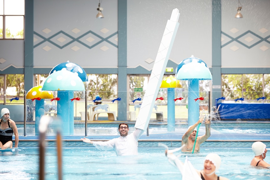 A man in white in an indoor pool, a long white structure extending from his arm. Four other people in the pool wear white caps