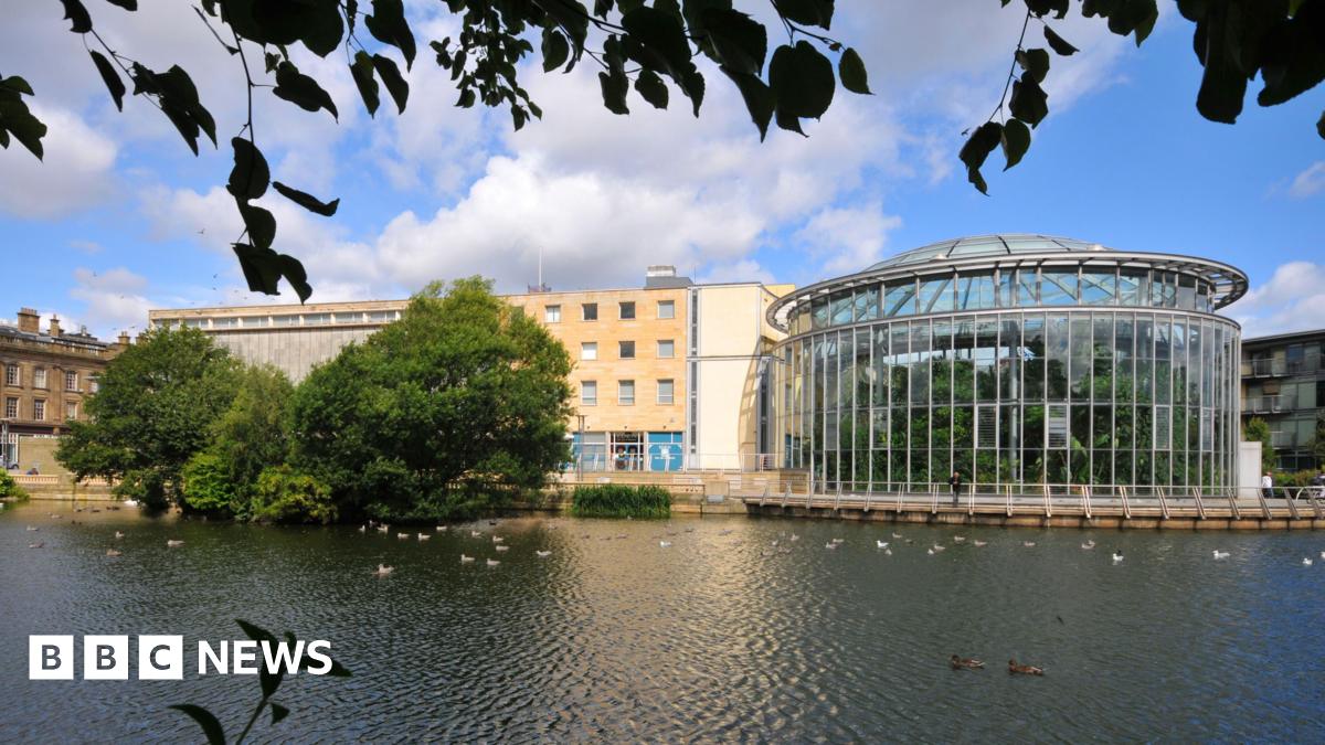 Brick building with multiple windows and  curved glass building. There are trees in front of it and a large pond.