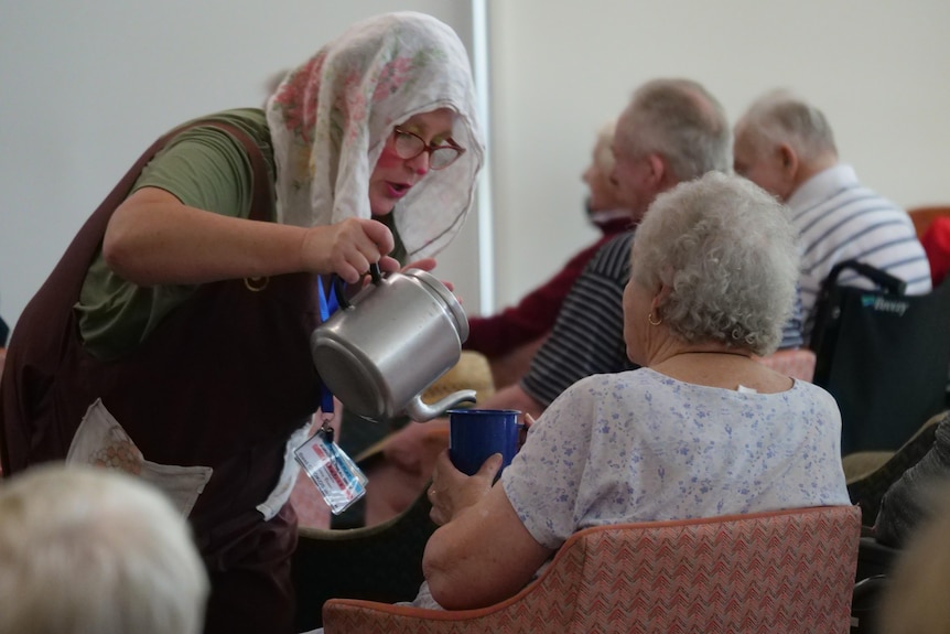 A woman holding a prop teapot pretends to pour tea for an elderly audience member.